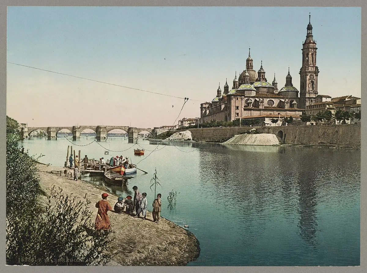 A tinted old photograph of the domes and towers of the Basilica of Our Lady of the Pillar in Zaragoza rising beyond the Ebro River, with people on the riverbank