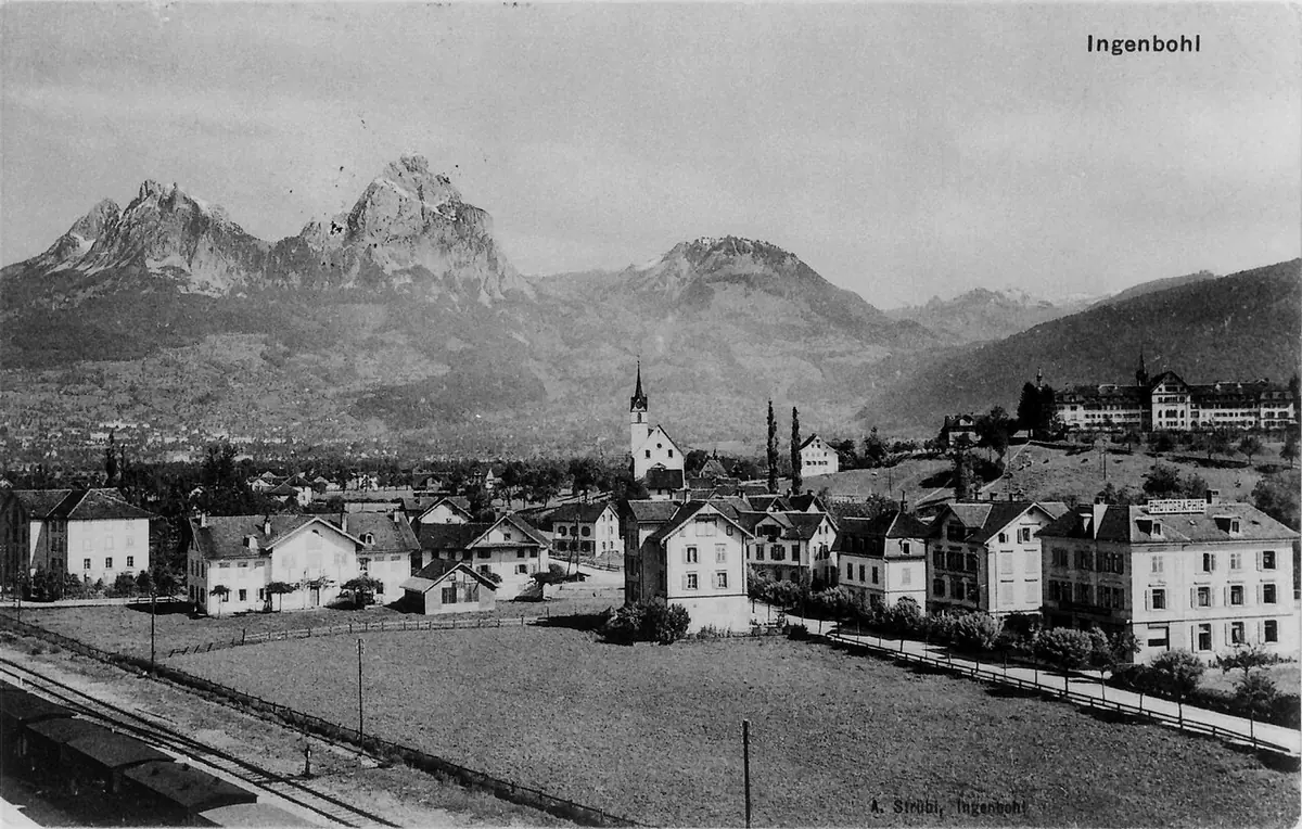 Old photograph of a village with a church spire and large convent buildings on a slope, beneath two sharp rocky mountain peaks