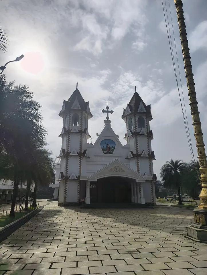 White church with two tall pointed towers and a cross over the entrance, seen across a paved courtyard with palm trees under a cloudy Kerala sky