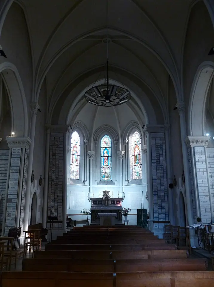 White vaulted nave of a small chapel with rows of wooden pews leading to an altar under three tall stained-glass windows