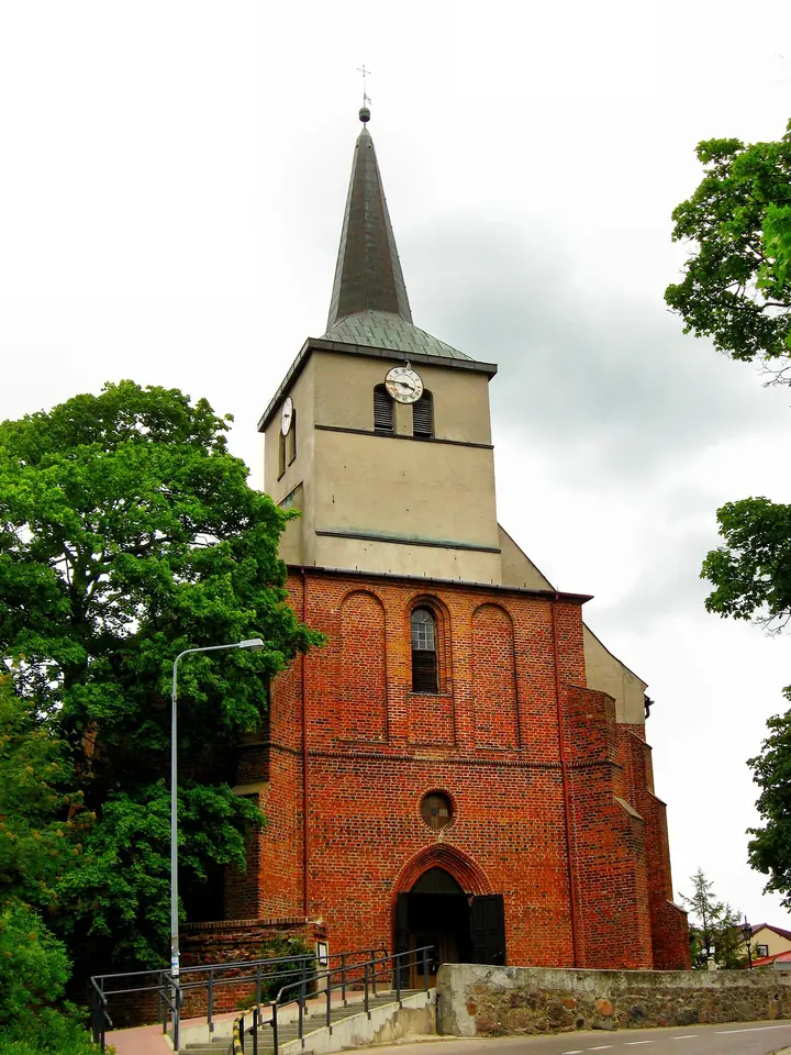 Photograph of a red-brick Gothic church with a tall square tower and a green spire among trees