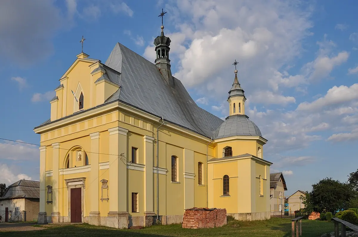 Photograph of a yellow Baroque-style Catholic church with a gabled front, a small domed side chapel and a slender lantern tower under a blue sky