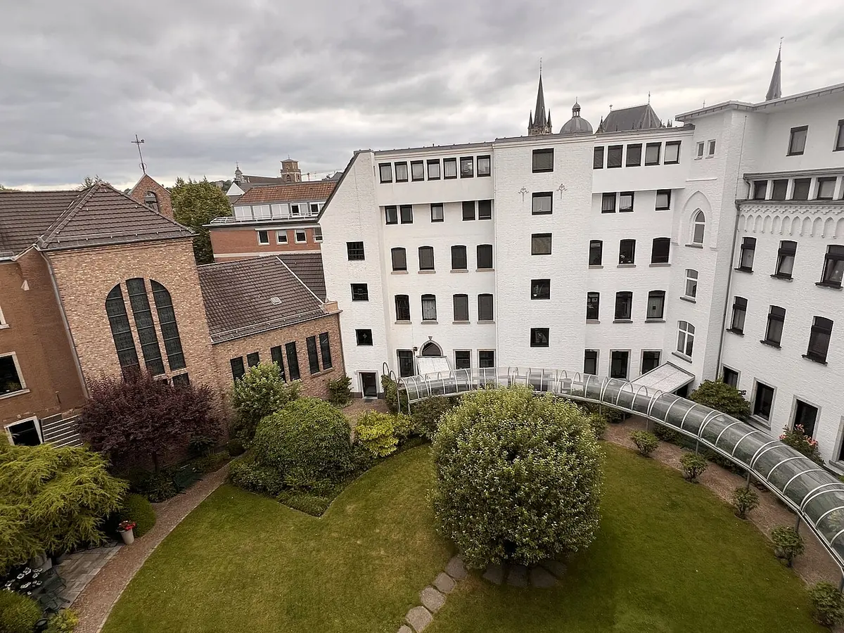 A green inner courtyard with a round lawn and shrubs, surrounded by white and brick convent buildings, with church spires in the distance