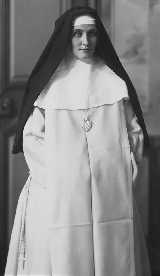 Old black-and-white photograph of a tall nun standing in a white habit and long black veil, with a heart-shaped emblem on her breast and a rosary at her side