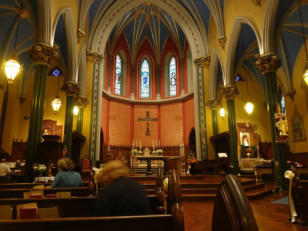 Interior of a Gothic church with green columns, painted ribbed vaults, pews and a sanctuary with a crucifix and tall stained-glass windows