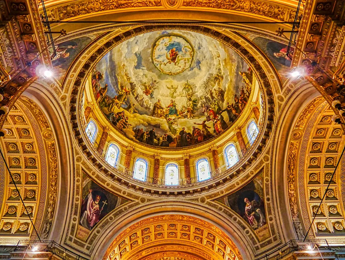View up into a gilded church dome painted with a crowd of saints and angels around the Virgin Mary, ringed with arched windows