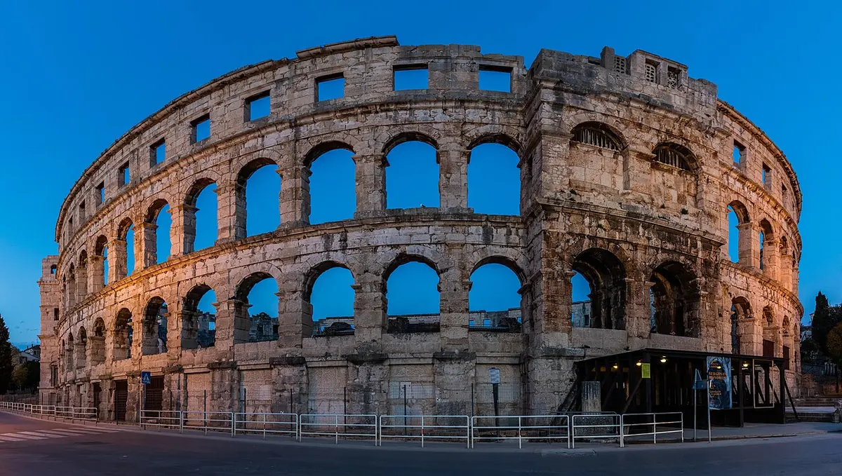 The Roman arena of Pula with its two tiers of stone arches, lit warmly at dusk against a deep blue sky