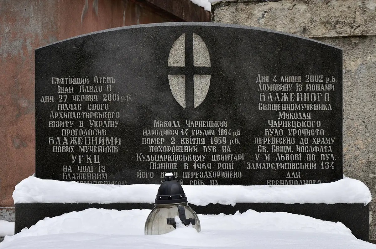 A black granite memorial stone with a cross and Ukrainian inscriptions, half covered in snow, with a grave lantern in front
