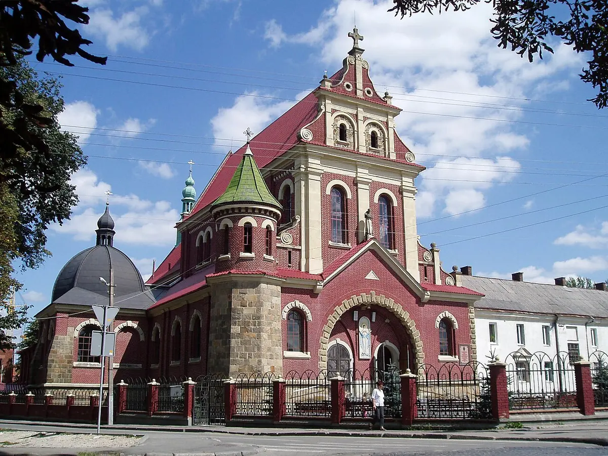 A large red-brick church with cream stone trim, a pointed central gable, a green dome and slender corner towers, behind an iron fence on a sunny day