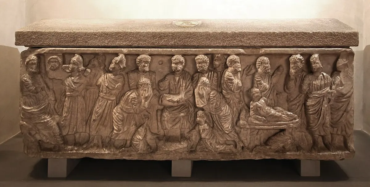 Ancient carved stone sarcophagus with a row of standing robed figures in relief along its front, in a softly lit crypt