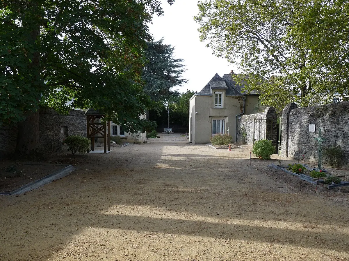 A quiet gravel lane between low stone walls and trees leading to a small stone house at the Champ des Martyrs in Avrillé