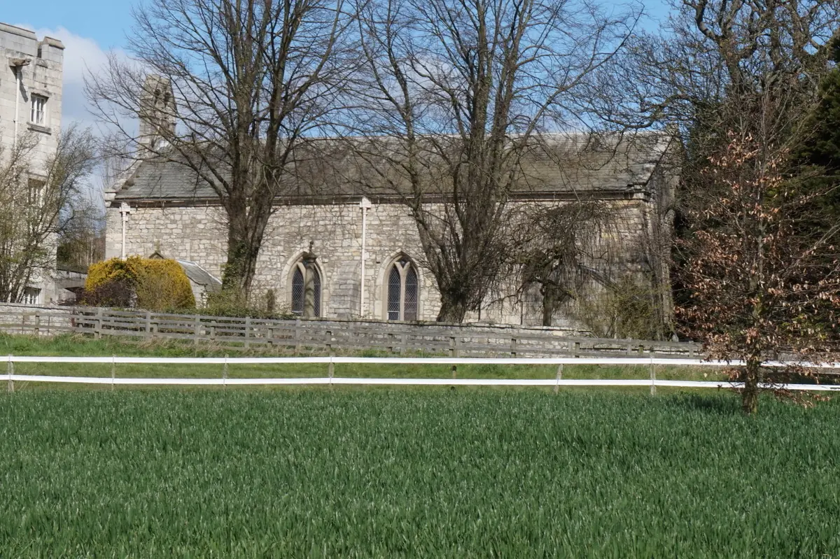 A small medieval stone chapel with pointed windows beside bare trees and a green field at Hazlewood Castle in Yorkshire