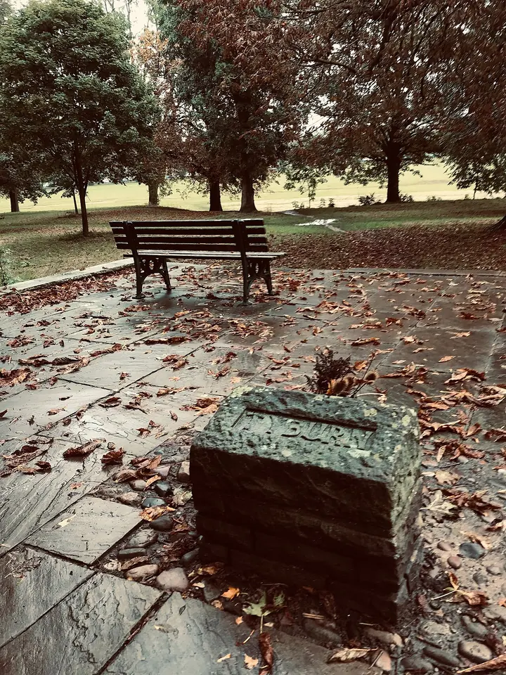 A low stone marker among fallen autumn leaves beside a bench and trees at the edge of the Knavesmire in York, the site of the city's old gallows