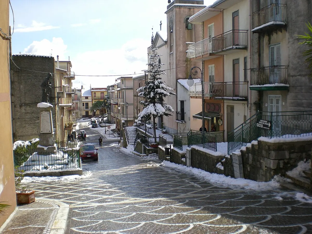 A sloping paved street in the center of Godrano lined with houses and balconies, with patches of snow on the steps and a small tree dusted with snow