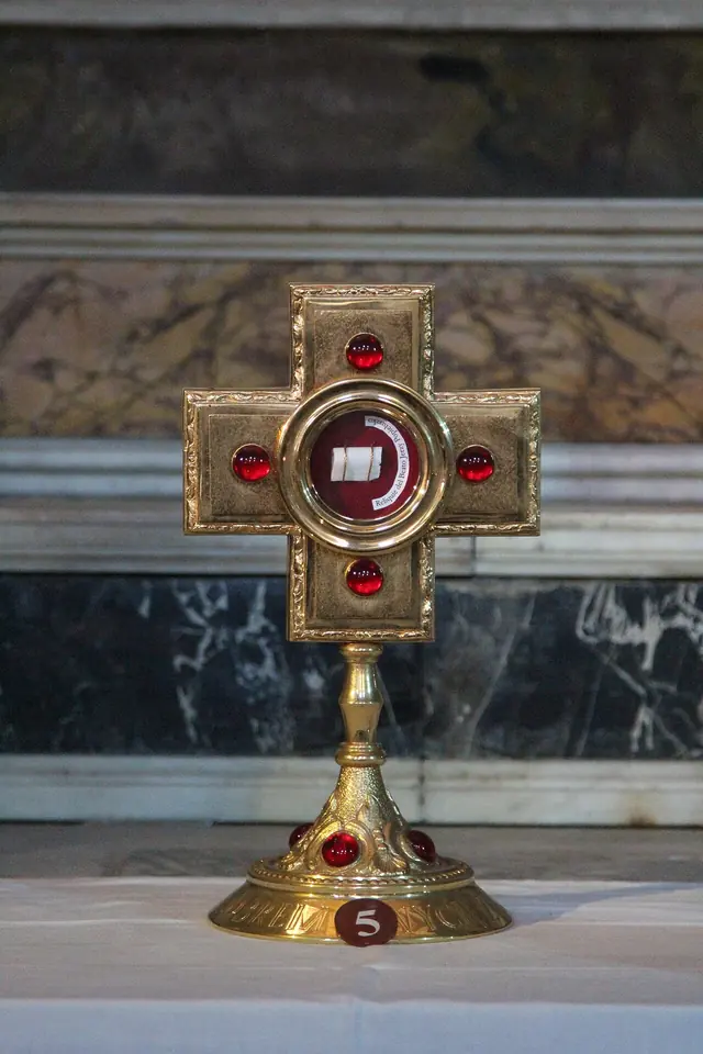 A gilded cross-shaped reliquary set with red stones and holding a small relic, standing on a marble altar