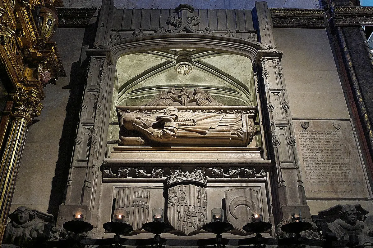 Gothic stone wall tomb with a carved recumbent figure under a pointed arch, set in a dim chapel with candles below