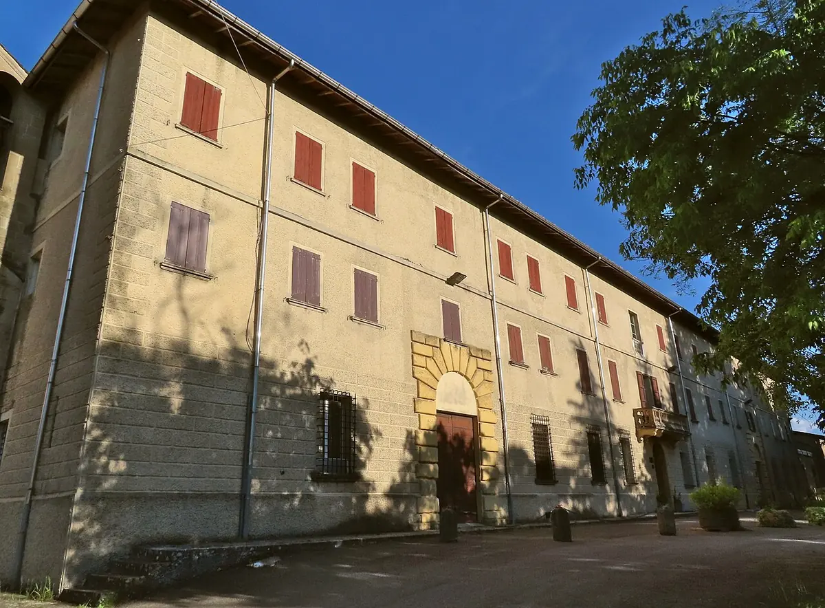 A long three-story yellow building with red shutters and an arched stone doorway, beside a tree, under a blue sky
