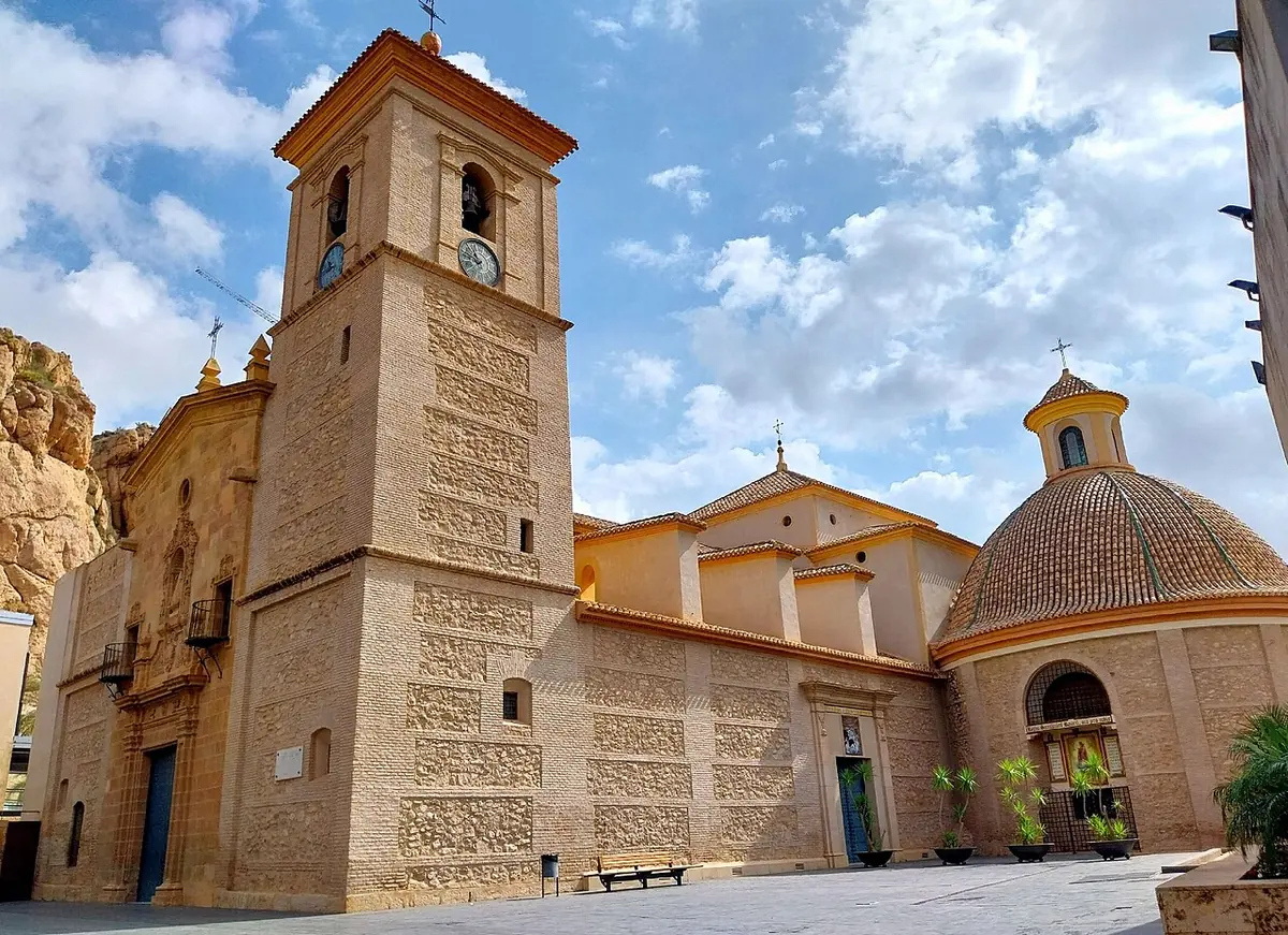 A golden stone church with a square bell tower, a carved doorway and a tiled dome beneath a rocky hill