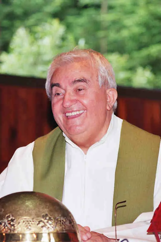Photograph of a smiling elderly priest with white hair, wearing a white alb and a green stole, seated behind a golden chalice