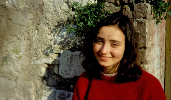Photograph of a smiling young woman with long dark hair in a red sweater, standing against an old stone wall with ivy