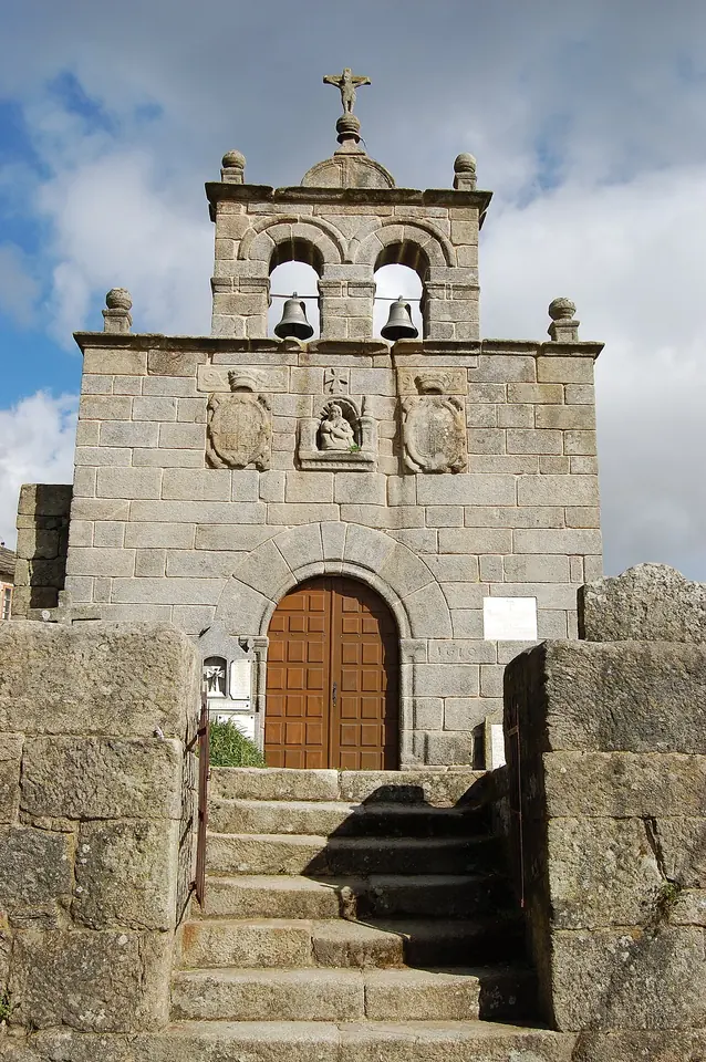 Small granite village church with a double bell gable, a carved coat of arms over an arched wooden door and stone steps leading up to it