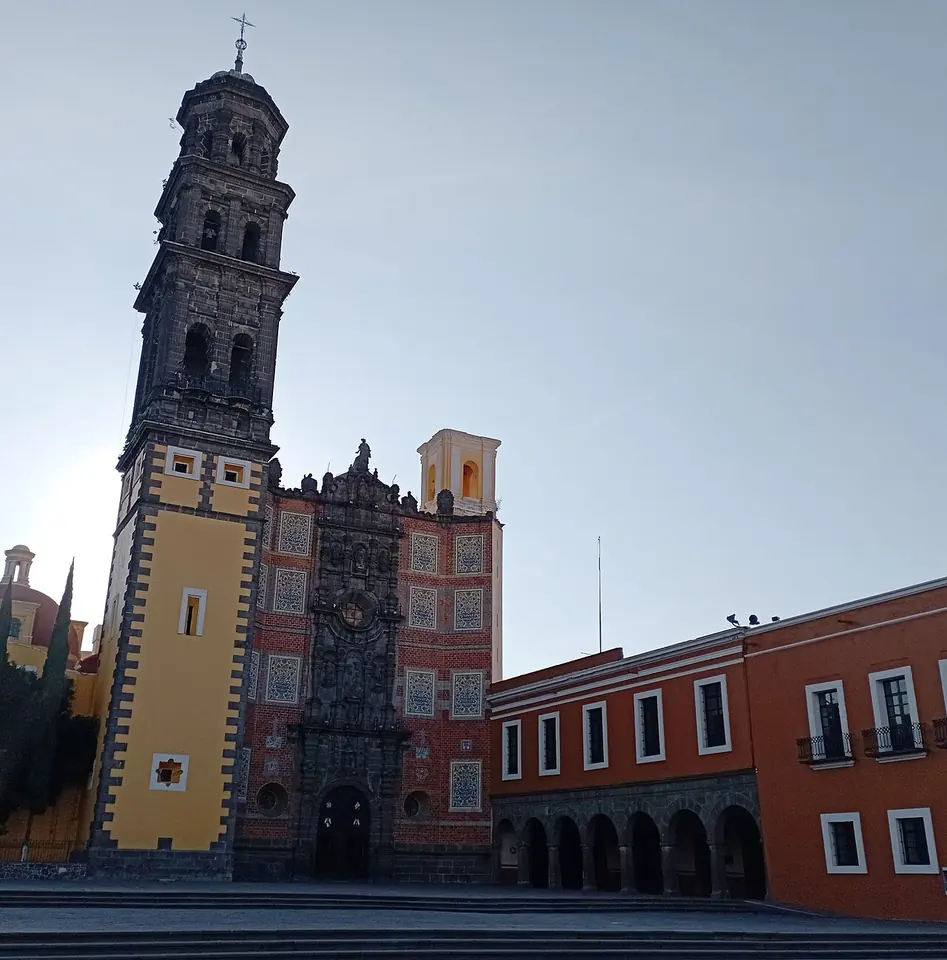 Tall dark stone bell tower beside a red-tiled Baroque church facade and the long orange convent building in Puebla, Mexico