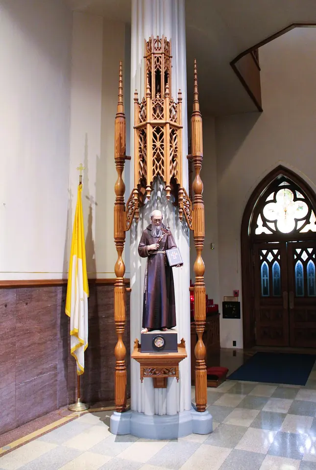 A painted statue of a bearded friar in a brown habit in a carved wooden Gothic niche beside a papal flag