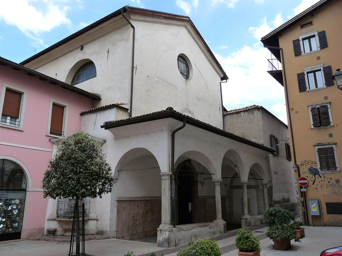 The white front of the old church of San Marco in Trento with an arcaded porch, between pink and yellow town houses