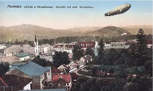 Old colored postcard view over the roofs of a small town with church towers and green hills, an airship in the sky, and a caption in Hungarian and German along the top