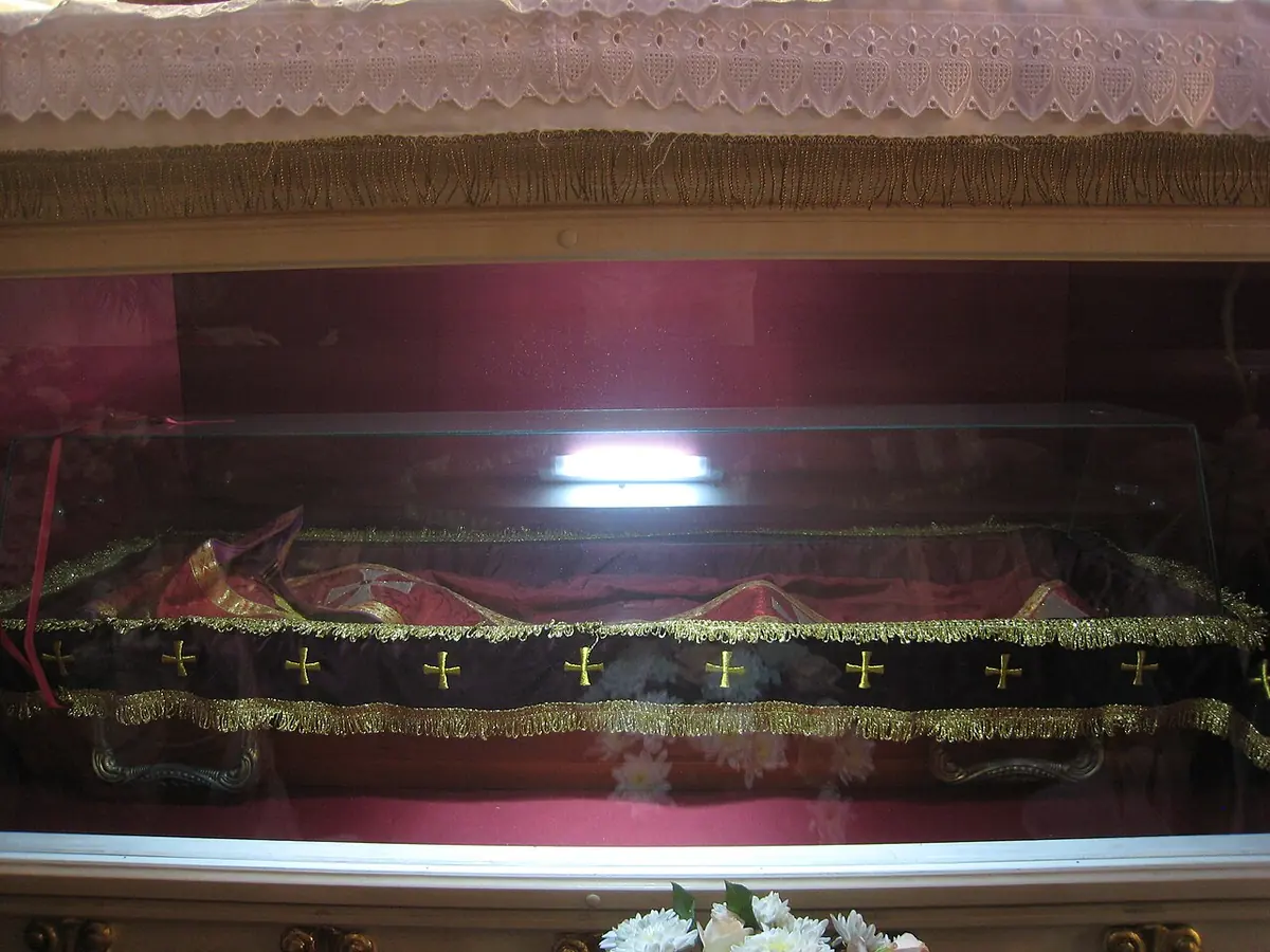 A glass reliquary chest draped in red and black cloth with small gold crosses, under a lace-edged canopy