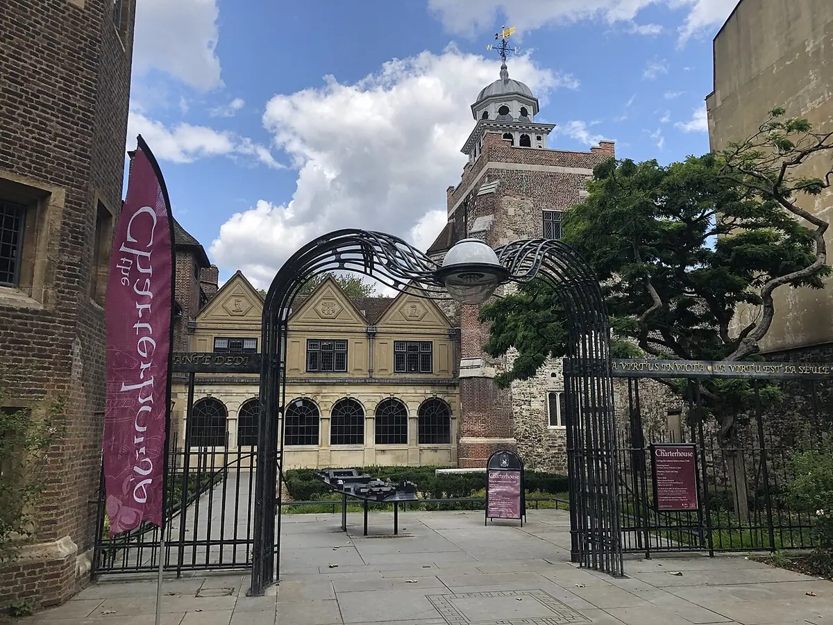 Wrought-iron gates opening onto a paved courtyard with old brick and stone buildings and a small cupola tower under a blue sky