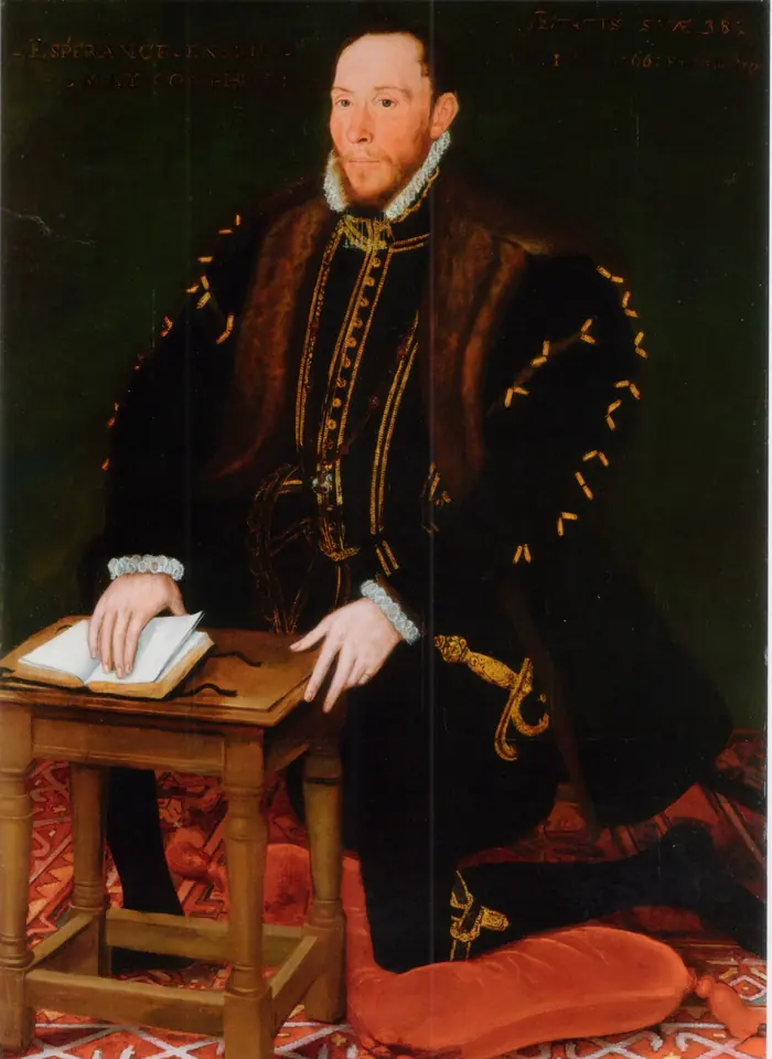 Full-length painted portrait of a bearded Tudor nobleman in black kneeling on a red cushion, wearing the Garter collar and reading an open prayer book on a small desk