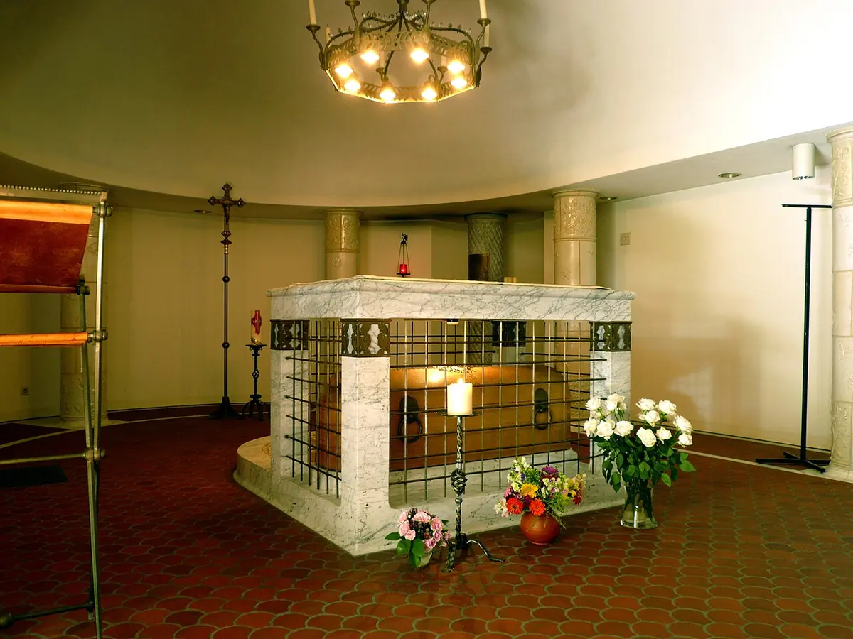 Low marble tomb enclosed by a metal grille in the middle of a round crypt, with flowers, candles and a crucifix