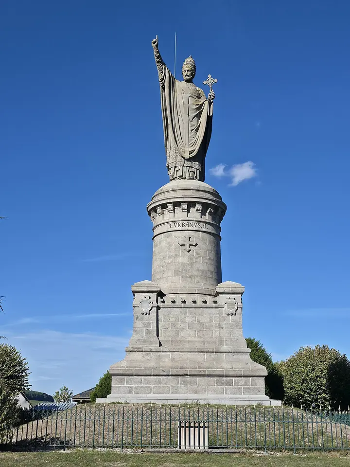 Colossal stone statue of a pope in a tiara holding a cross and raising one arm, on a tall round pedestal against a blue sky