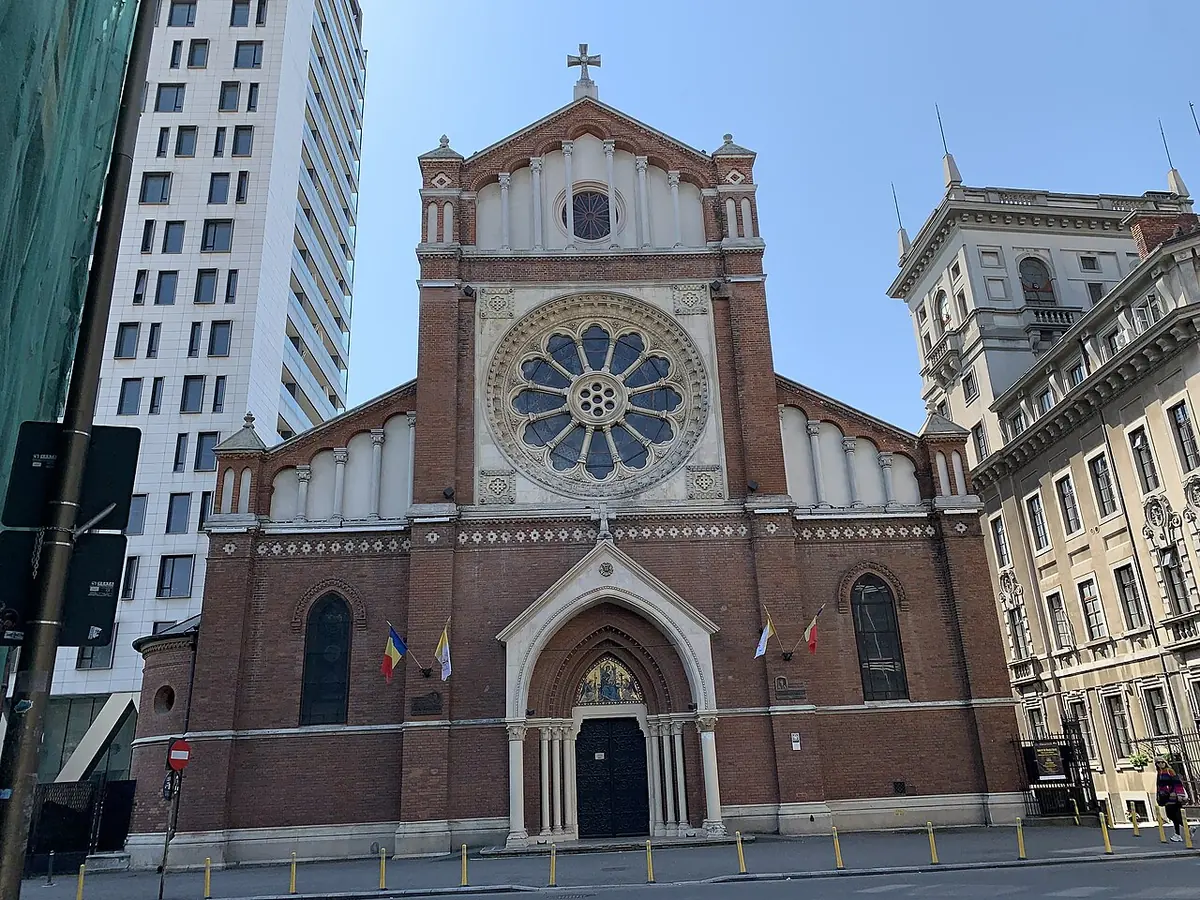 Red-brick neo-Romanesque cathedral front with a large rose window and an arched doorway, between modern buildings in Bucharest