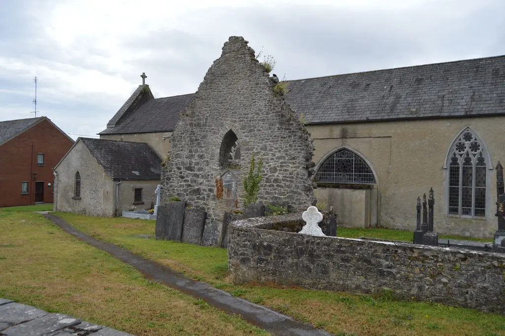 The roofless gable of a medieval stone friary with a tall pointed window beside a later stone church, with graves and a low wall in front