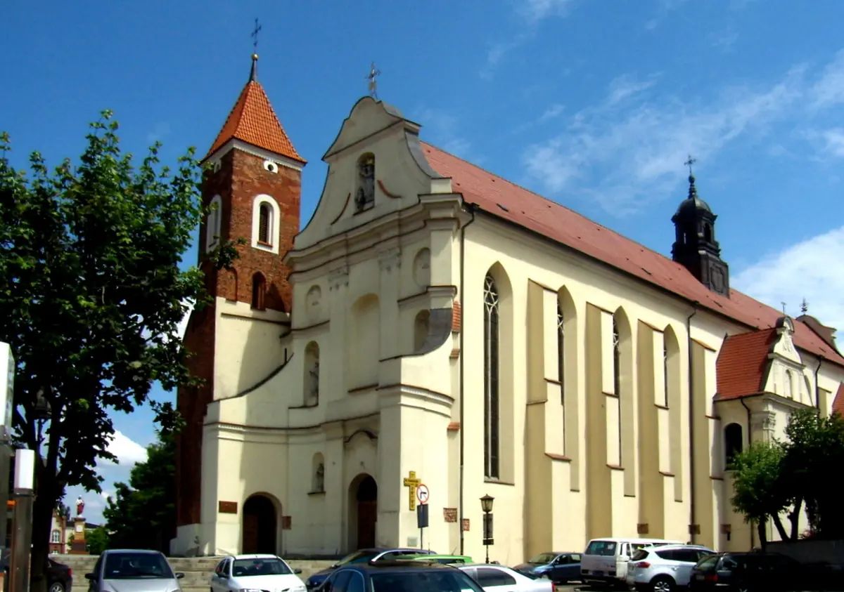 A long cream-colored church with a baroque front, a red-roofed brick bell tower and a small spire, under a blue sky with cars parked in front