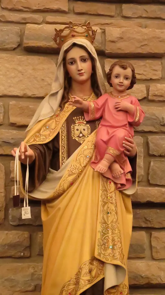 Painted statue of a crowned Mary in a brown tunic and cream mantle holding the Child Jesus in pink, with small scapulars hanging from her hand