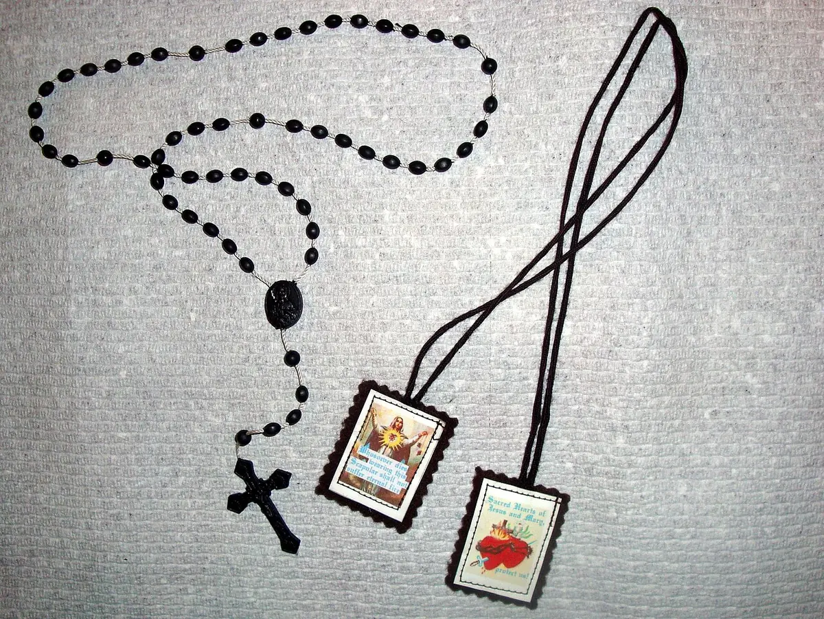 Black rosary beads and a brown scapular with two small picture squares, one of Our Lady of Mount Carmel and one of the Sacred Heart, laid on a gray cloth