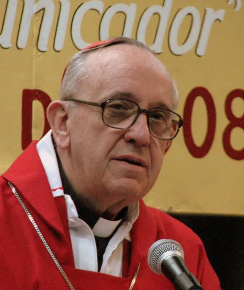 An older man with glasses in a red chasuble and red zucchetto speaking into a microphone, a banner in the background