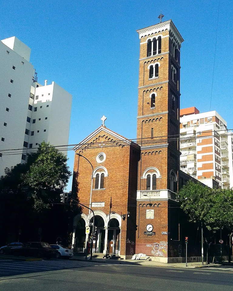 A red-brick Romanesque-revival church with an arched portico and a tall square bell tower topped by a cross, on a city avenue between apartment blocks under a blue sky