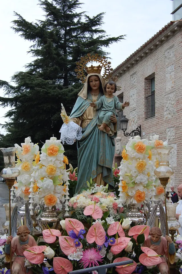 A statue of the Virgin holding the Christ Child, crowned and surrounded by white and pink flowers, carried on a float through a town street