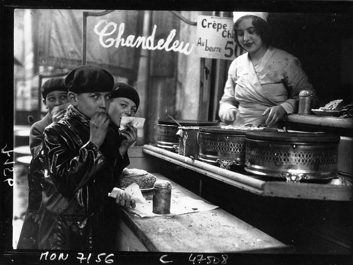 Black-and-white press photo of boys in caps eating crêpes at a street stall with a sign reading Chandeleur, while a woman in a white apron cooks
