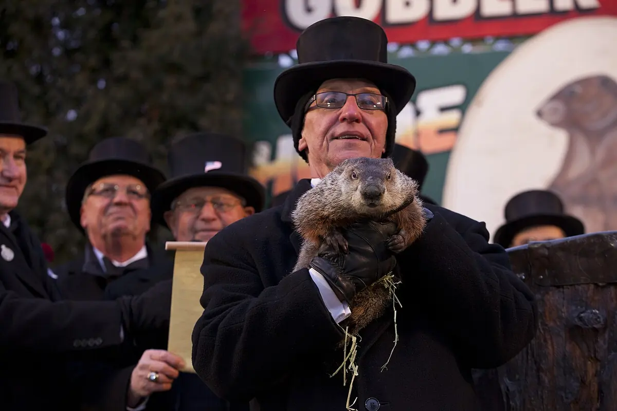A man in a black top hat holds up a groundhog in front of a crowd of other men in top hats on a stage