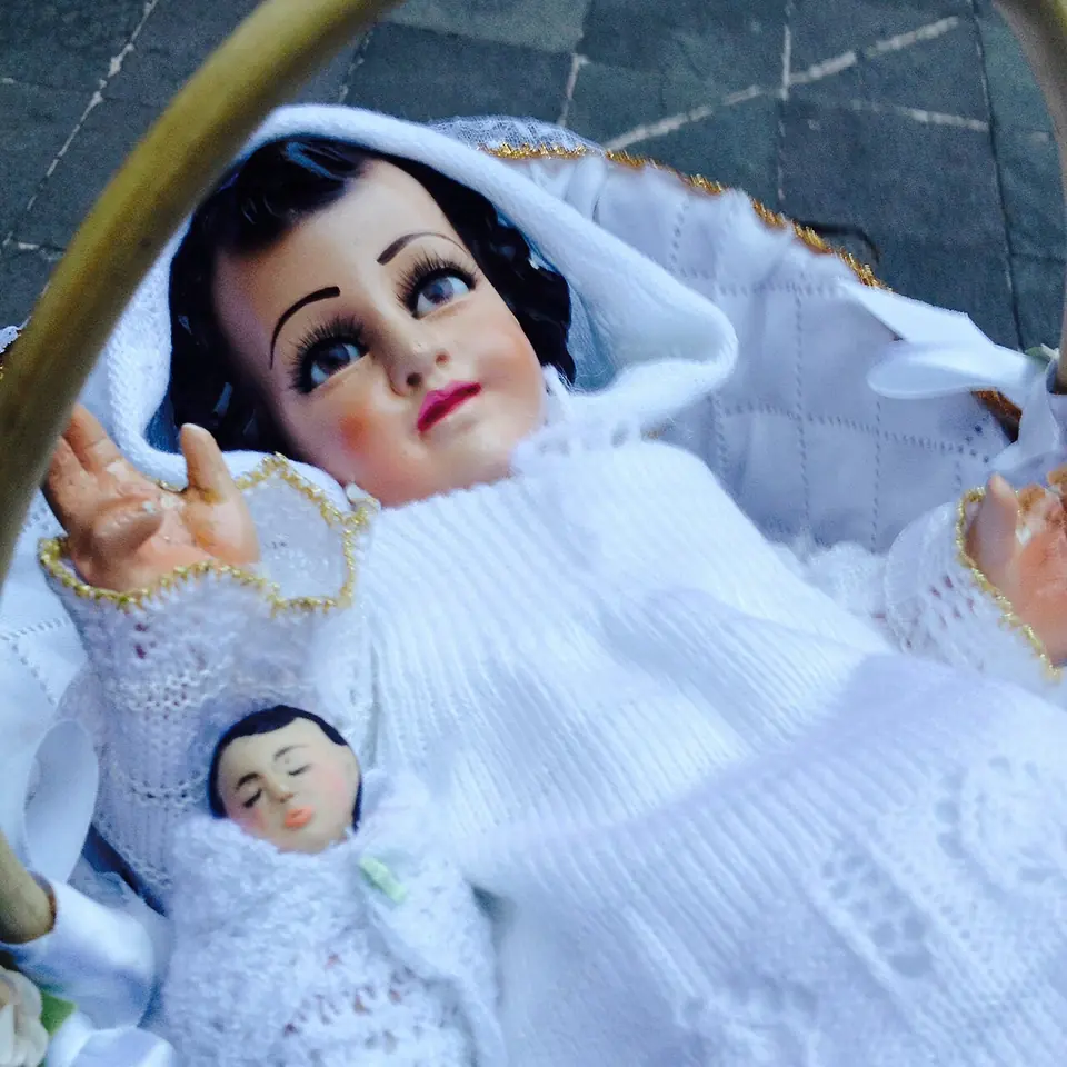 A doll-like statue of the infant Jesus in a white lace gown and hood lying in a basket, with a smaller dressed figure of the Christ Child beside it