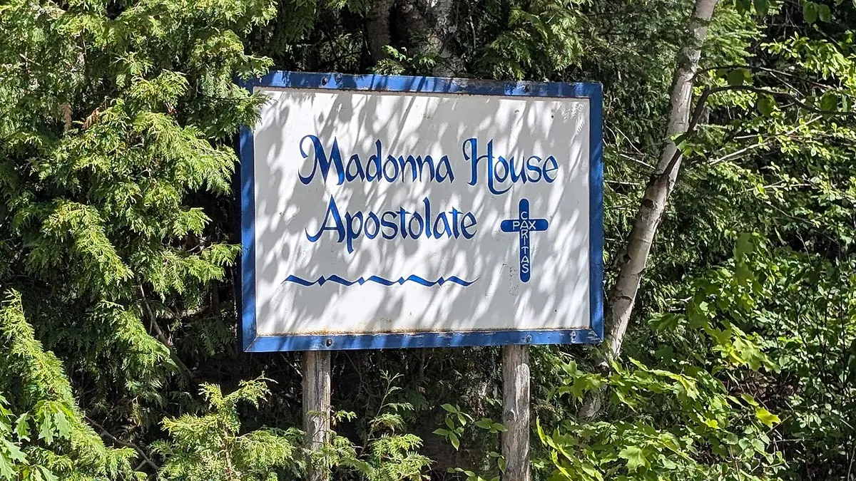 A white wooden sign with blue lettering reading Madonna House Apostolate and a small cross, among green cedar branches