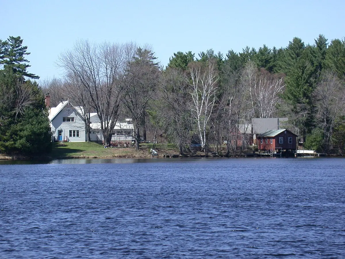White wooden houses and a red boathouse among spring trees on the far bank of a wide blue river