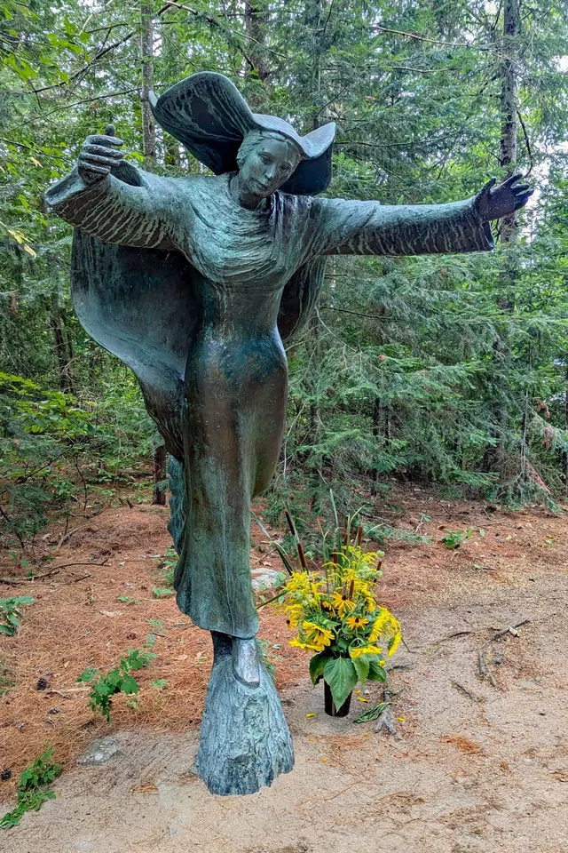 Bronze statue of the Virgin Mary running forward with her arms stretched wide and her veil blowing behind her, among pine trees, with yellow flowers at her feet