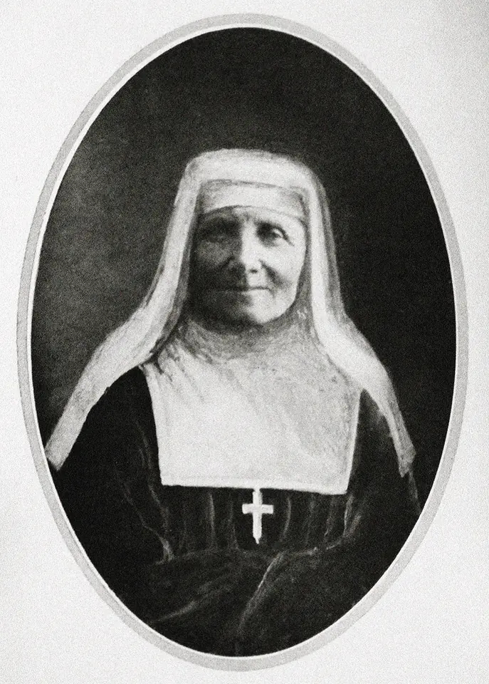 Oval photograph of an elderly smiling nun in a black habit, a white wimple and a black veil, with a small white cross on her chest