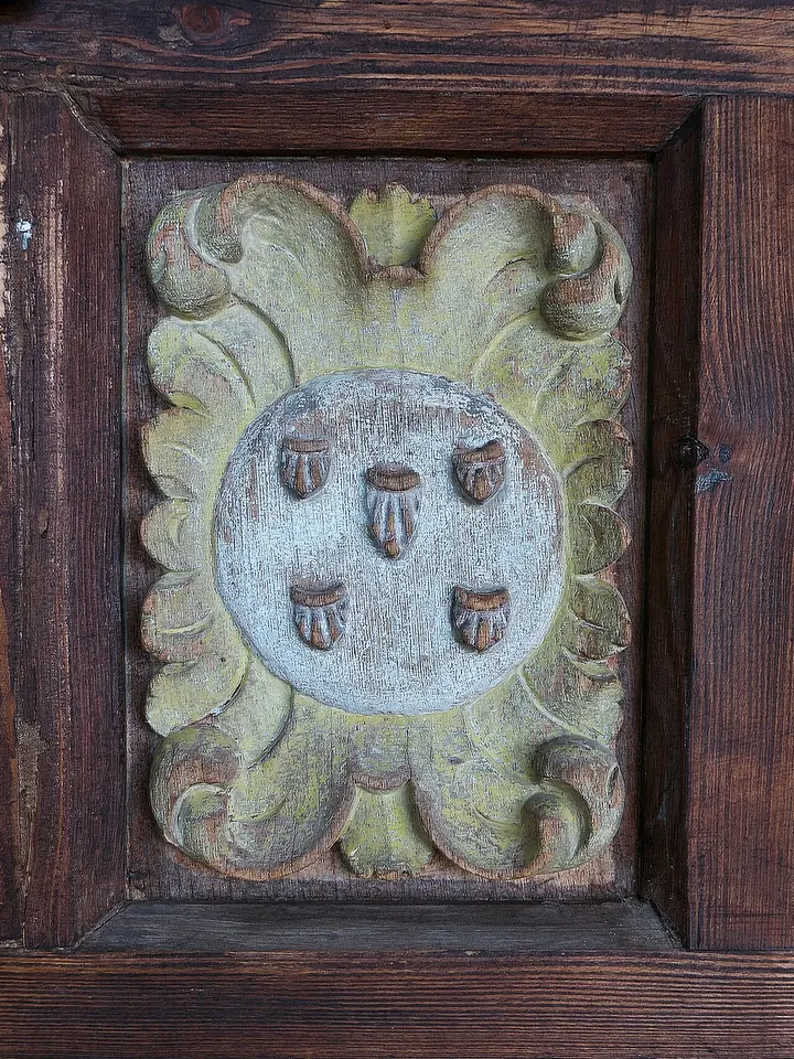 Carved and painted wooden door panel with a silver medallion showing the five wounds of Christ arranged in a cross pattern inside a scrolled green frame